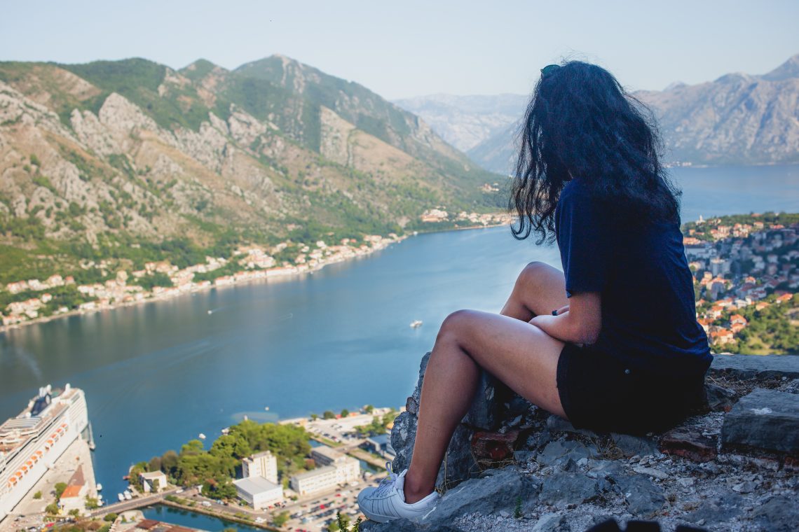 A woman with curly dark hair sits on a rocky ledge overlooking the Bay of Kotor in Montenegro. She wears a navy shirt, black shorts, and white sneakers, gazing out at the sweeping view of the bay, surrounding mountains, and distant town below.